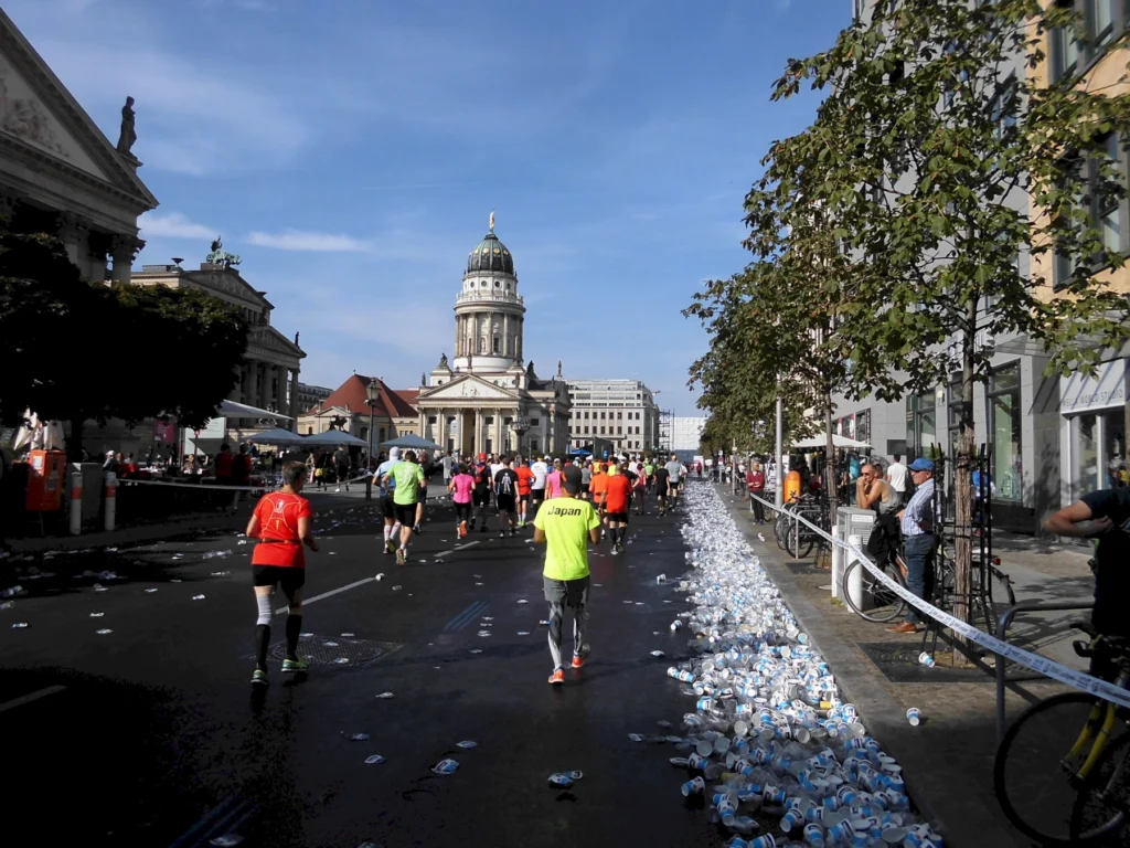 Läufer beim Marathon in einer Stadtstraße, umgeben von verstreuten Trinkbechern und historischen Gebäuden im Hintergrund, unter blauem Himmel.