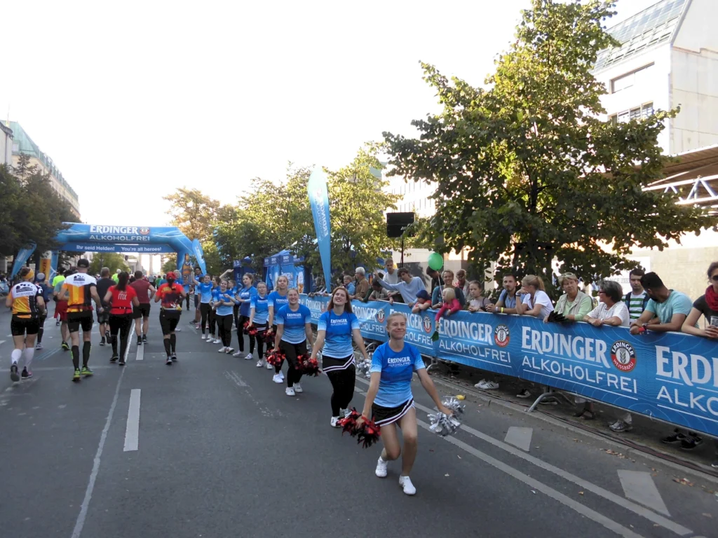 Eine Gruppe von Cheerleadern in blauen T-Shirts feuert Läufer bei einem Marathon an, während Zuschauer an der Strecke stehen. Im Hintergrund ist ein Banner der Marke Erdinger zu sehen, das für alkoholfreies Bier wirbt.
