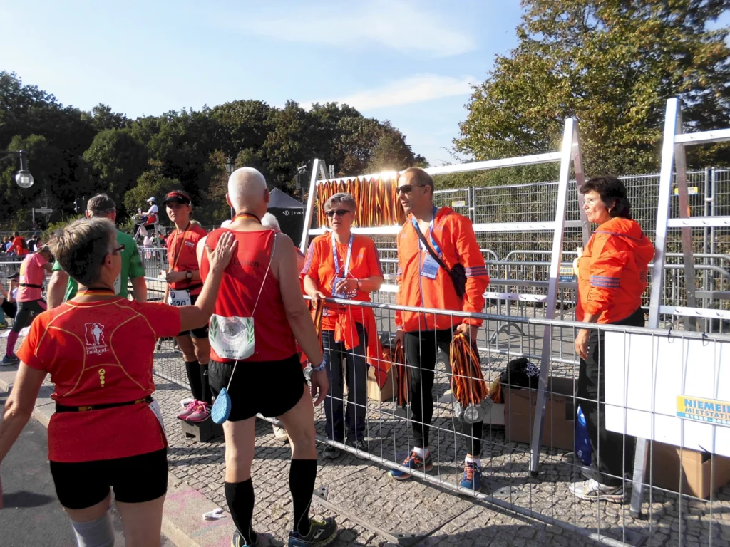 Teilnehmer eines Laufwettbewerbs in roten Trikots stehen an einem Absperrgitter, während orange gekleidete Helfer Informationen und Medaillen verteilen. Im Hintergrund sind weitere Läufer und eine festliche Atmosphäre zu sehen.