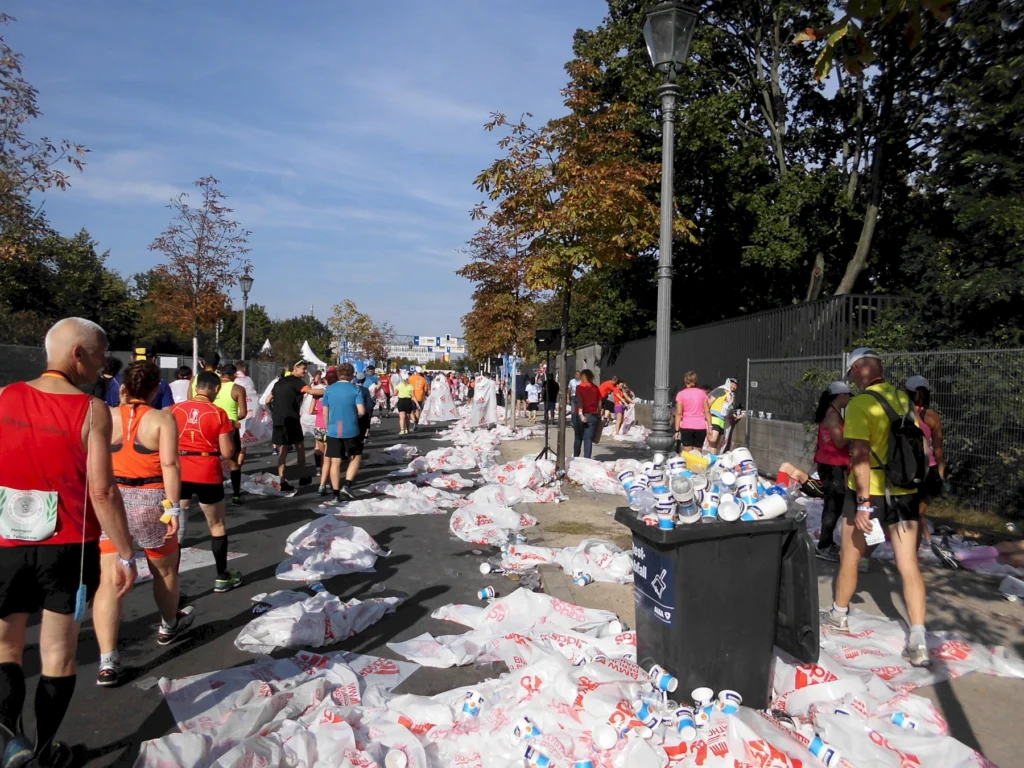 Läufer auf einer Straße nach einem Marathon, umgeben von leeren Trinkbechern und Müllsäcken, sonniger Tag, Herbstbäume.