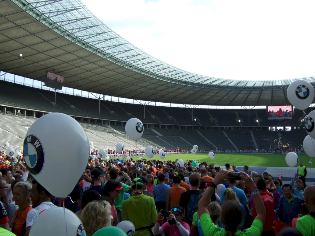 Menschenmenge im Stadion mit BMW-Luftballons, während Teilnehmer an einem Sportevent warten. Im Hintergrund ist eine große Leinwand zu sehen, die das Geschehen im Stadion zeigt.