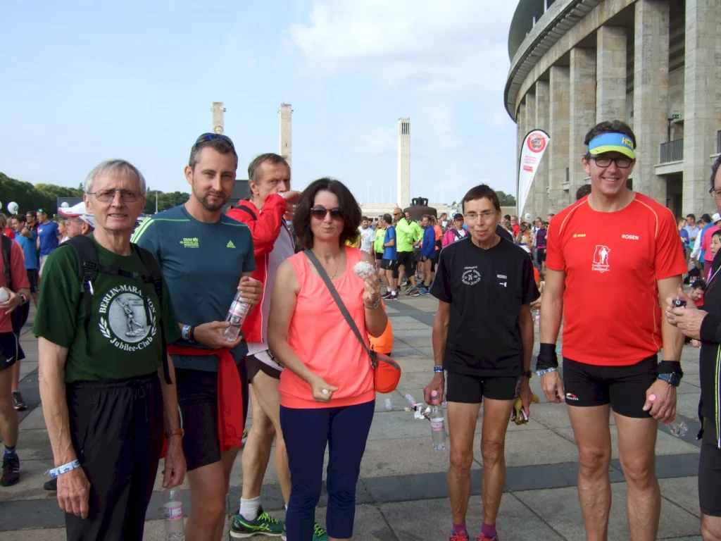 Gruppe von Läufern in sportlicher Kleidung beim Berlin-Marathon, stehen vor dem Olympiastadion. Einige halten Wasserflaschen, während sie auf den Start warten.