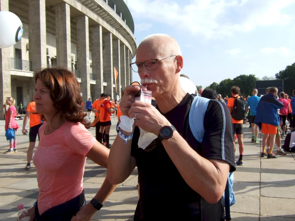 Älterer Mann trinkt bei einem Lauf-Event vor dem Stadion Berlin, umgeben von Teilnehmern in bunter Sportkleidung.
