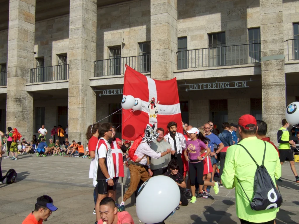 Gruppierung von Teilnehmern bei einem Lauf-Event vor einem historischen Gebäude, mit einer großen Flagge und Luftballons. Menschen in Sportkleidung stehen zusammen, einige lächeln und posieren für Fotos.