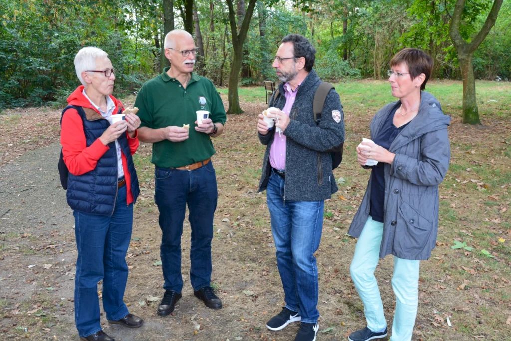 Gruppe von vier Personen unterhält sich im Park mit Getränken in der Hand, umgeben von herbstlicher Natur.