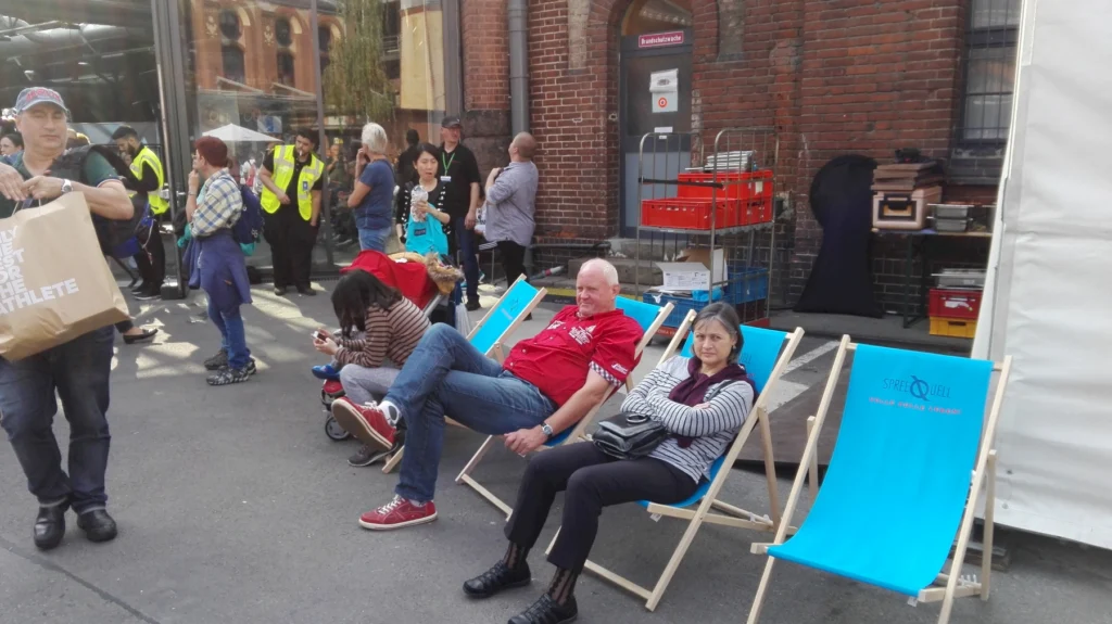 Menschen entspannen sich in blauen Liegestühlen vor einem Backsteingebäude auf einer belebten Straßenszene.