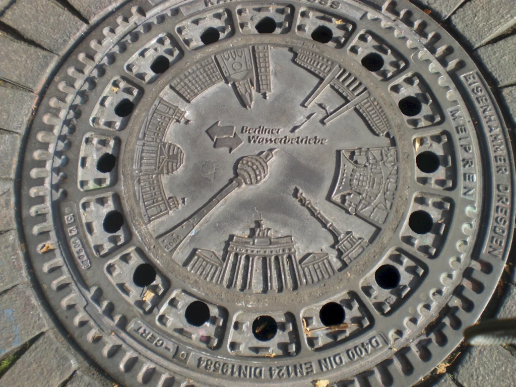 Gullydeckel in Berlin mit eingeprägten Wahrzeichen der Stadt wie dem Fernsehturm und Brandenburger Tor.