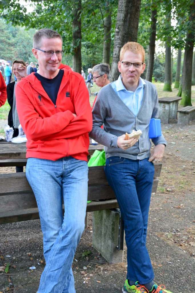 Zwei Männer stehen entspannt an einer Bank im Freien. Der Mann links trägt einen roten Hoodie und hat die Arme verschränkt, während der Mann rechts ein Sandwich hält und in einem grauen Cardigan gekleidet ist. Im Hintergrund sind weitere Personen und Bäume zu sehen, was eine gesellige Atmosphäre vermittelt.