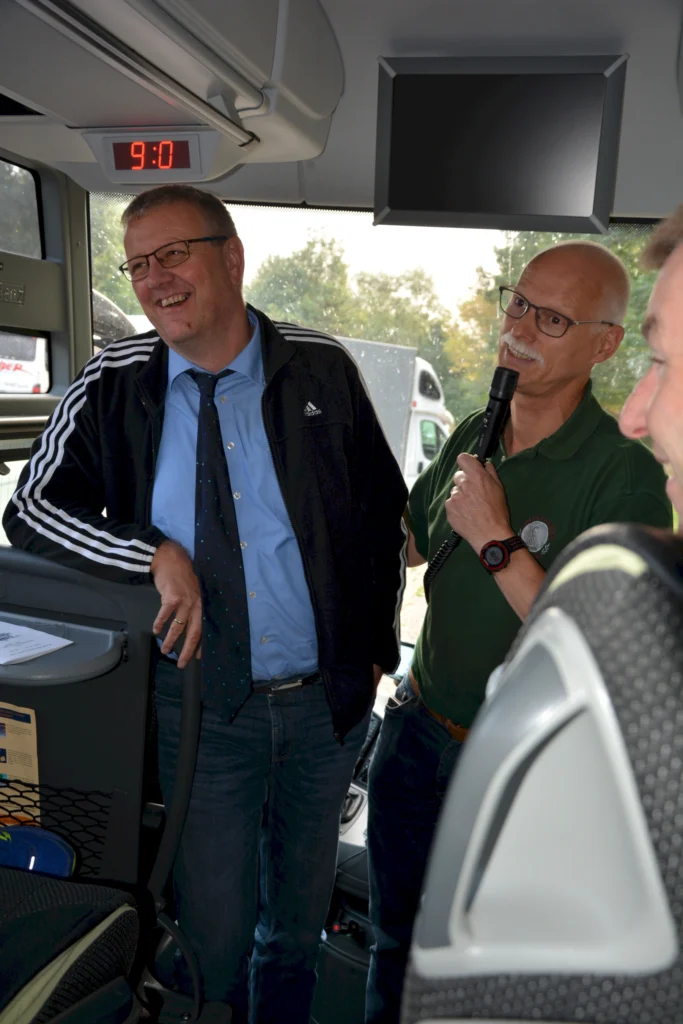 Zwei Männer unterhalten sich fröhlich in einem Bus, einer hält ein Mikrofon in der Hand.
