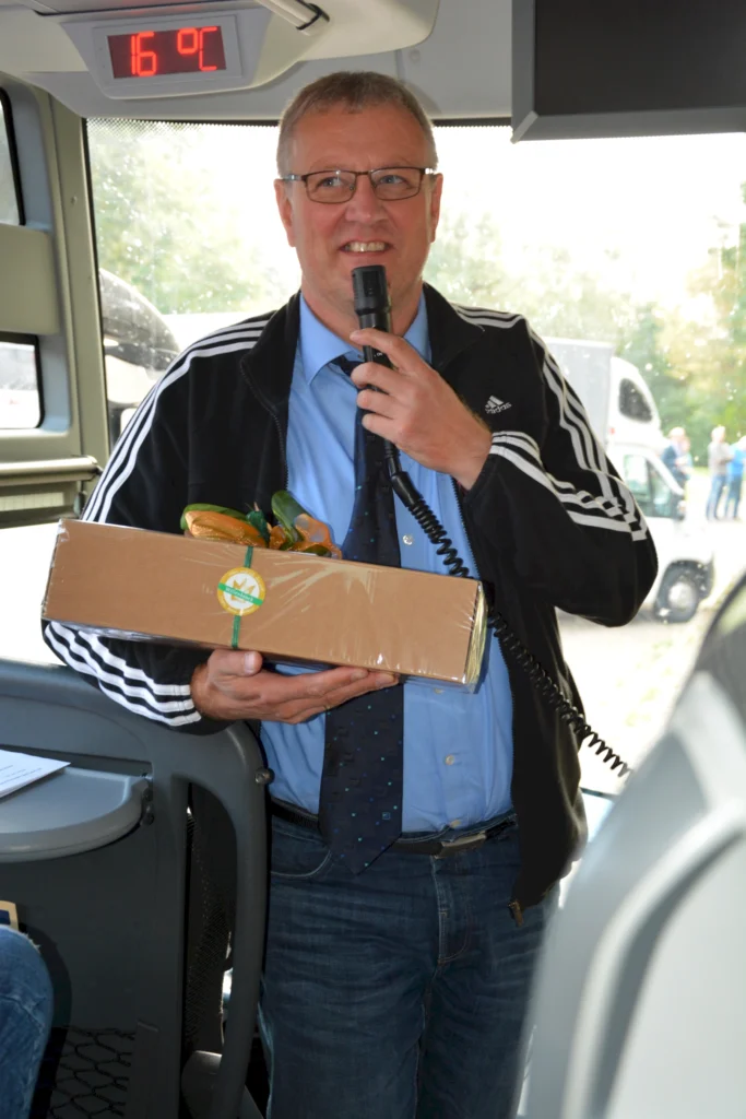 Mann in Bus mit Mikrofon und Geschenk, spricht zu Passagieren. Temperaturanzeige zeigt 16 °C an.