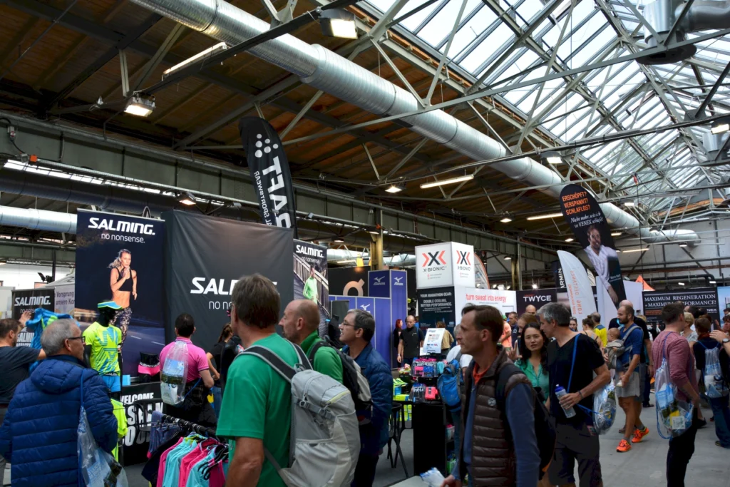 Messestand auf einer Sportmesse mit Besuchern, die sich über Sportbekleidung und -zubehör informieren. Marken wie Salming und X-Bionic sind prominent vertreten. Die Atmosphäre ist lebhaft und interaktiv, mit einer Vielzahl von Produkten und Werbematerialien.