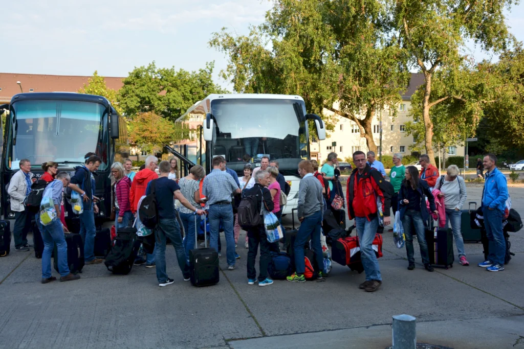 Gruppe reisender Menschen mit Gepäck vor Bussen, bereit zur Abfahrt, bei sonnigem Wetter auf einer Straße.