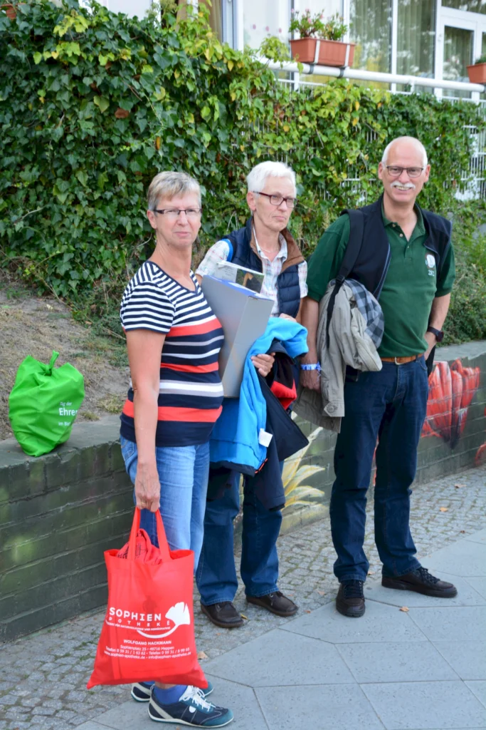 Drei Menschen mit Taschen stehen vor einer mit Efeu bewachsenen Mauer. Sie tragen Freizeitkleidung.