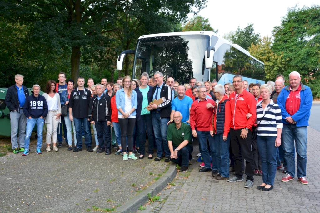 Gruppe von Menschen vor Reisebus im Freien, umgeben von grünen Bäumen, bereitet sich auf eine gemeinsame Fahrt vor.