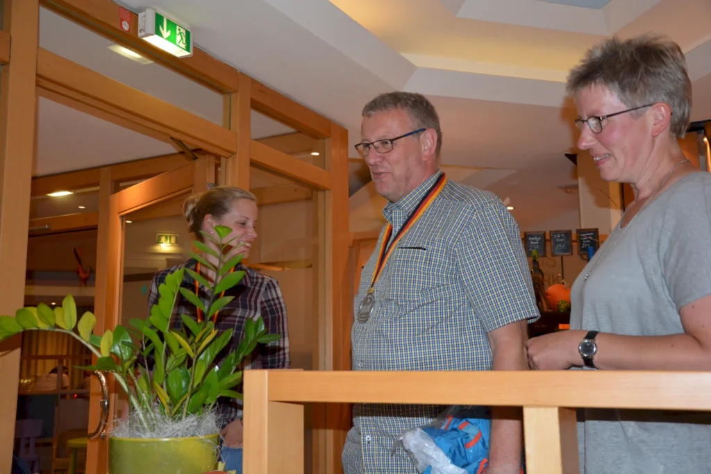 Drei Personen stehen in einem Restaurant, während eine Frau lächelnd hinter einer Pflanze steht. Ein Mann in einem karierten Hemd trägt eine Medaille und hält eine Tüte, während eine weitere Frau neben ihm steht. Die Umgebung ist hell und einladend, mit einer Holzverkleidung und Dekorationen im Hintergrund.