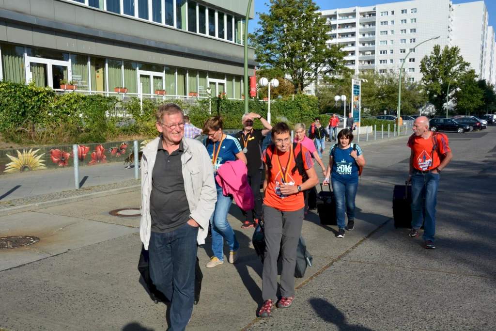 Gruppe von Personen mit Gepäck geht lächelnd auf einem sonnigen Bürgersteig neben einem Gebäude mit Pflanzen.