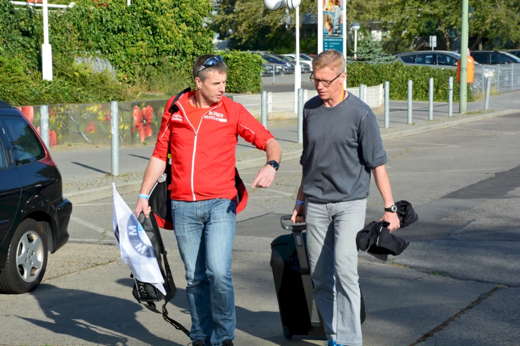 Zwei Männer mit Gepäck und Sportbekleidung auf Straße, diskutierend. Umgebung: Autos, Natur und Gebäude im Hintergrund.