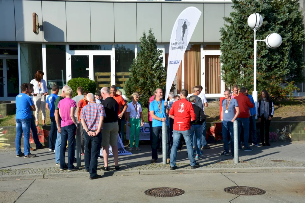 Gruppe von Menschen bei einem Sportevent vor einem Gebäude, einige tragen Medaillen, sonniges Wetter, Eventfahne sichtbar.