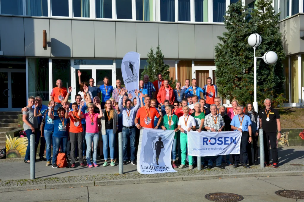 Gruppe fröhlicher Menschen mit Medaillen und Bannern vor einem Gebäude, lächelnd und winkend bei sonnigem Wetter.