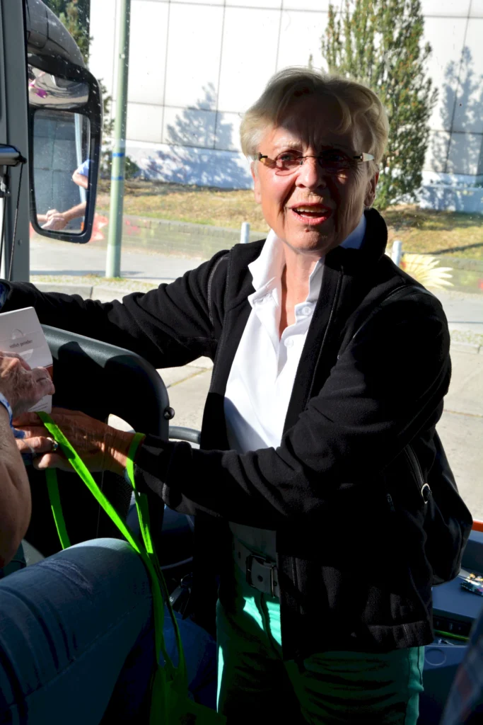 Ältere Frau mit Brille lächelt in einem Bus, trägt eine weiße Bluse und schwarze Jacke. Fröhliche Atmosphäre.