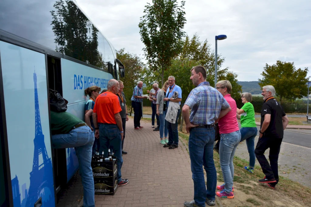 Gruppe von Menschen, die an einem Reisebus stehen, während sie sich unterhalten und Getränke entladen. Der Bus hat eine grafische Darstellung des Eiffelturms und trägt den Schriftzug „DAS HIGHLIGHT HIER“. Die Szene spielt sich an einem sonnigen Tag mit leicht bewölktem Himmel ab.