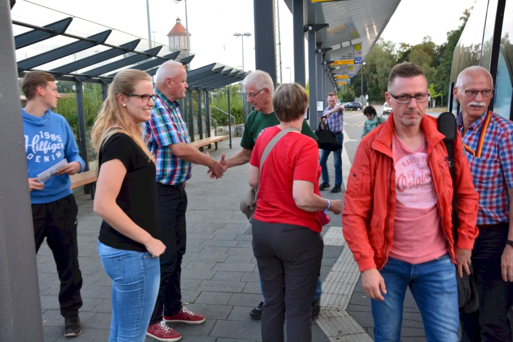 Menschen begrüßen sich und unterhalten sich an einer überdachten Bushaltestelle im Freien.