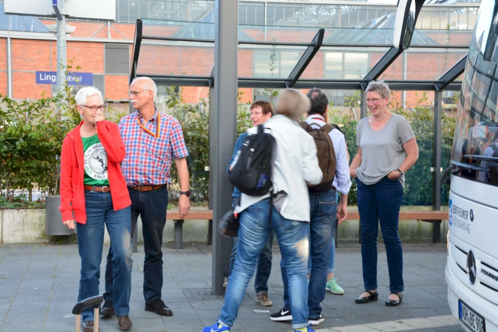 Gruppe von Menschen unterhält sich lachend an einer Bushaltestelle in einer städtischen Umgebung mit Glasüberdachung.