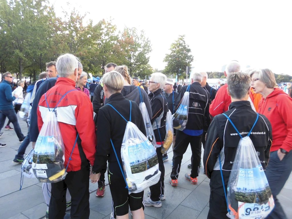 Gruppe von Läufern mit Startnummern und Taschen versammelt sich vor einem Rennen im Freien, umgeben von Bäumen.