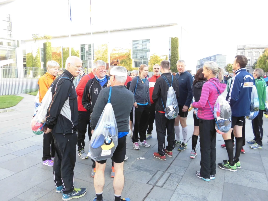 Gruppe von Läufern in Sportkleidung, die sich vor einer modernen Gebäudeansicht versammeln, während sie sich auf ein bevorstehendes Rennen vorbereiten. Einige Teilnehmer halten Tüten mit Laufsachen.
