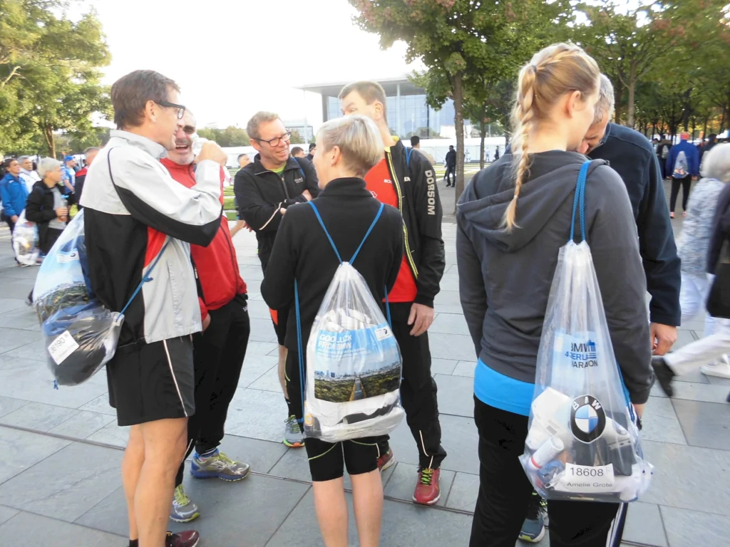 Gruppe von Läufern mit Sporttaschen beim Berlin-Marathon, plaudernd auf dem Sammelplatz vor dem Laufstart.