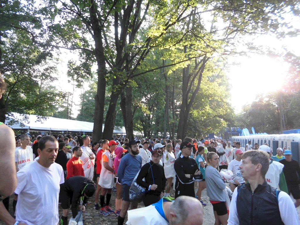 Teilnehmer eines Laufevents in Berlin versammeln sich in einem belebten Park, umgeben von Bäumen und einem Zelt. Die Atmosphäre ist lebhaft, mit Läufern in Sportkleidung, die auf den Start des Rennens warten.