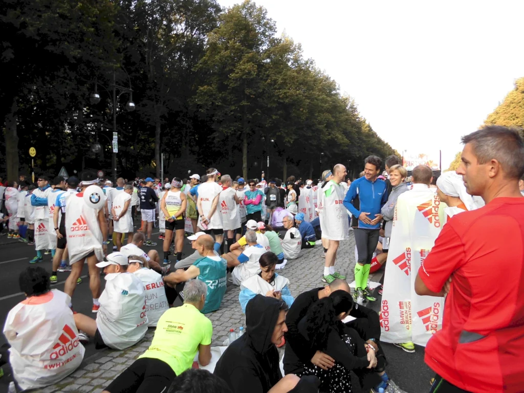 Teilnehmer eines Marathons sitzen und stehen in der Warteschlange, um am Rennen teilzunehmen. Viele tragen leichte Regenponchos mit dem Logo von Adidas. Die Szene spielt sich in einem urbanen Park mit Bäumen im Hintergrund ab.