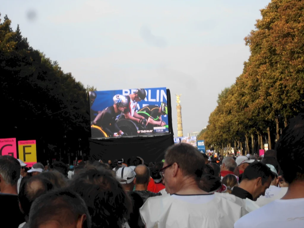 Menschenmenge bei einem Sportereignis in Berlin, mit großem Bildschirm im Hintergrund, auf dem Athleten zu sehen sind. Die Siegessäule ist in der Ferne sichtbar, umgeben von Bäumen und bunten Plakaten.