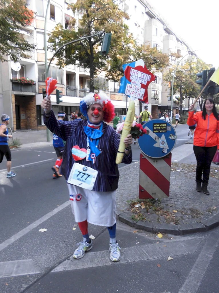 Läufer im Clownskostüm mit Motivationstafel bei Straßenmarathon, umgeben von Zuschauerinnen und Zuschauern in urbaner Kulisse.