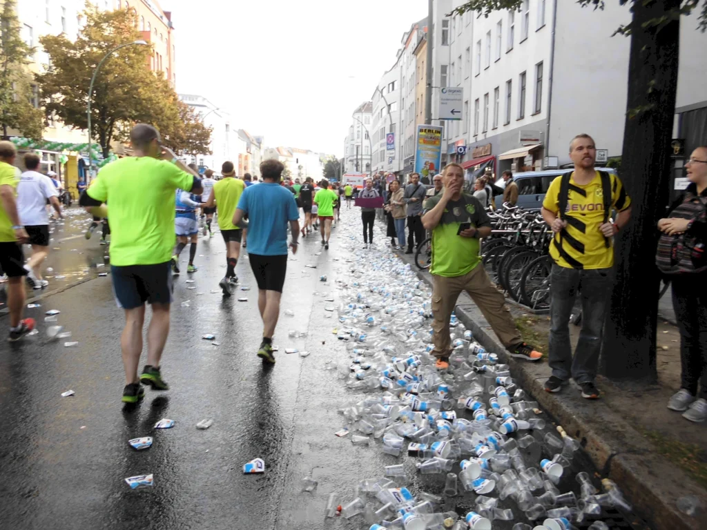 Läufer auf einer Straße mit Plastikbechern nach einem Rennen; Zuschauer am Rand stehen unter Bäumen.