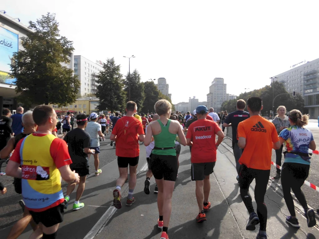 Läufer beim Marathon auf einer Stadtstraße, umgeben von Bäumen und Gebäuden, mit bunten Sportkleidungen und motivierten Gesichtern. Die Veranstaltung zieht zahlreiche Teilnehmer an, die sich in Richtung Ziel bewegen.