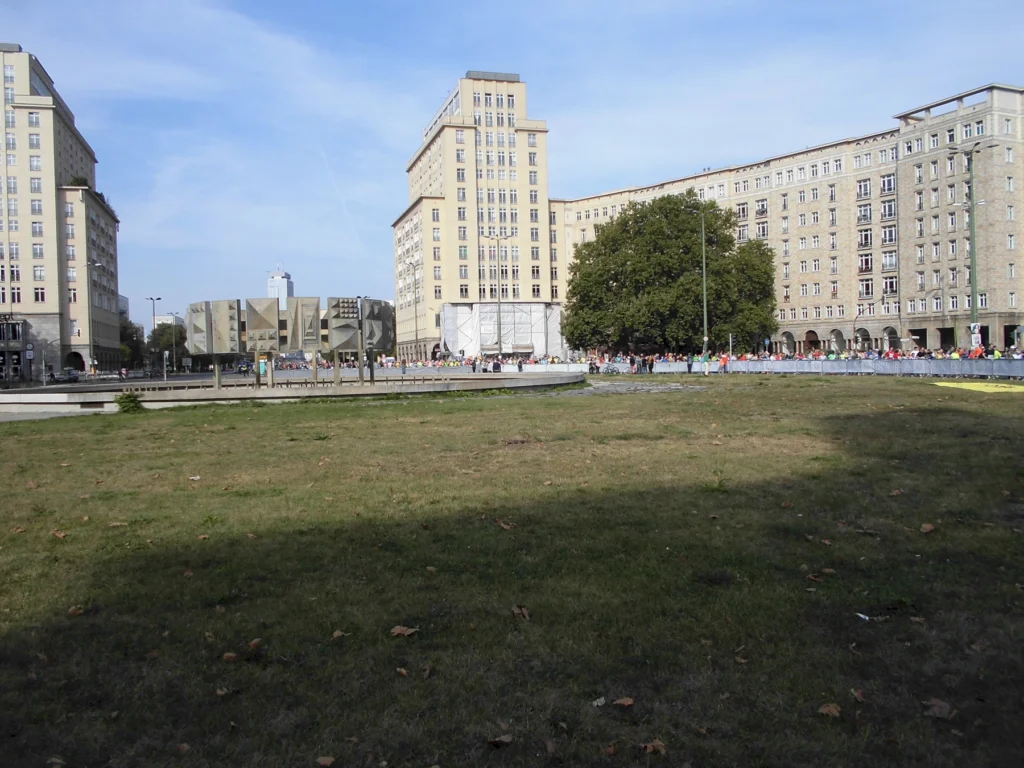 Grüne Wiese vor Gebäuden in einer Stadt mit Menschen im Hintergrund bei klarem Himmel.