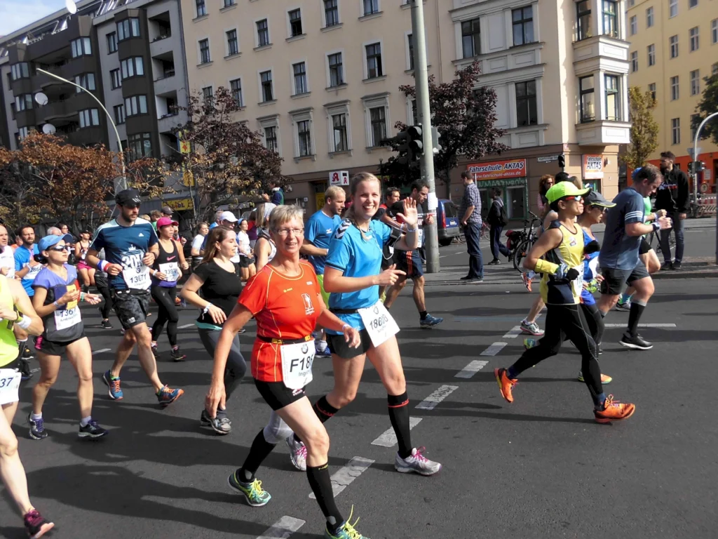 Läuferinnen und Läufer nehmen an einem Straßenrennen teil, während sie fröhlich durch die Stadt joggen. Die Teilnehmer tragen bunte Sportbekleidung und Wettkampfnummern. Im Hintergrund sind mehrstöckige Gebäude und Zuschauer zu sehen, die die Athleten anfeuern.