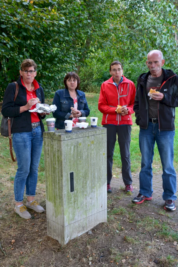 Vier Personen essen im Freien bei einem Ausflug. Sie stehen an einem bepflanzten Platz mit Kaffeebechern in der Hand.