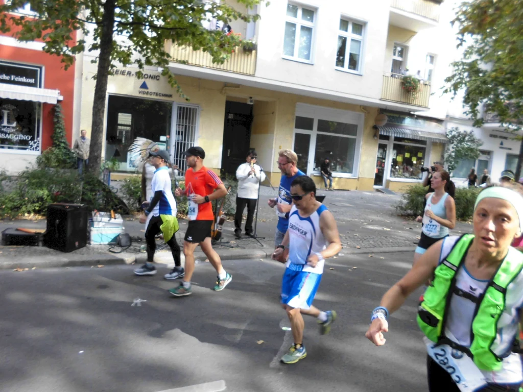 Läufer in verschiedenen Sportoutfits nehmen an einem Straßenrennen teil, während Zuschauer im Hintergrund die Veranstaltung beobachten. Die Szene zeigt eine lebendige Atmosphäre mit herbstlichen Bäumen und Geschäften im Hintergrund.