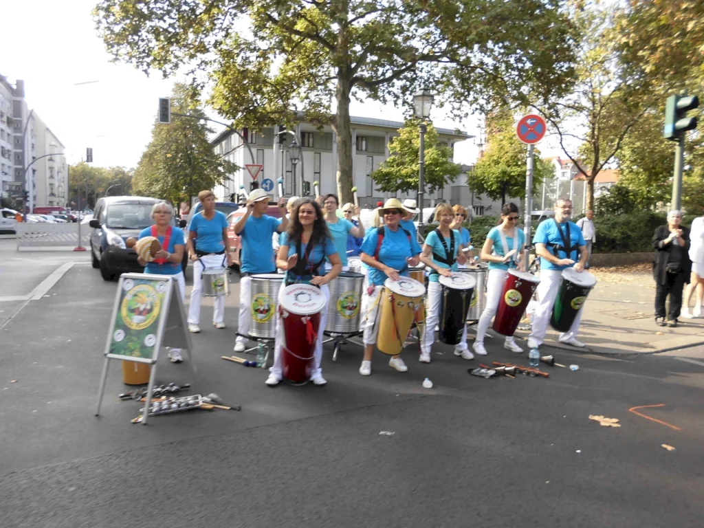 Straßenband in blauer Kleidung spielt Trommeln bei sonnigem Wetter auf einer belebten Kreuzung vor Publikum.