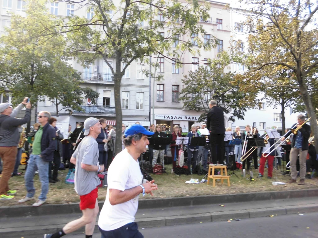Läufer und Blasorchester bei einer Outdoor-Veranstaltung in einer städtischen Umgebung mit Bäumen und einem Dirigenten.