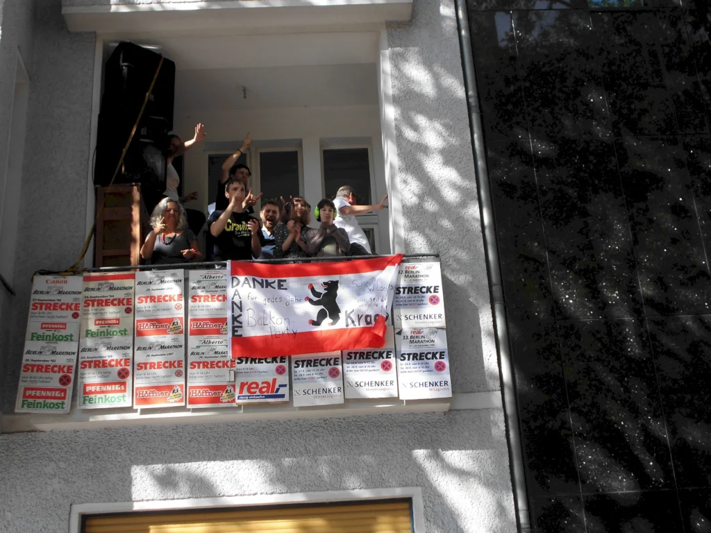Gruppierung von Menschen auf einem Balkon, die mit einer Flagge und Plakaten feiern. Die Flagge zeigt einen Bären und den Schriftzug "Danke für jedes Jahr Balkon Party". Im Hintergrund sind mehrere Werbeplakate sichtbar.