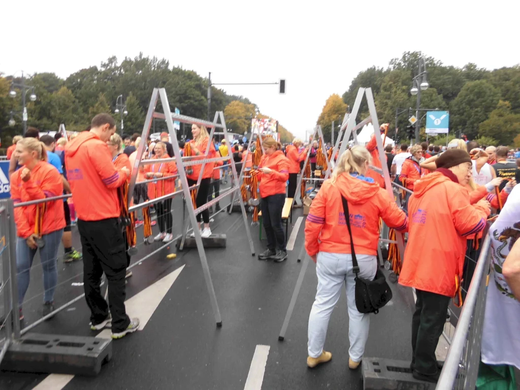 Menschen in orangefarbenen Jacken verteilen Medaillen an Marathonläufer auf einer Straße vor einer grünen Baumkulisse.