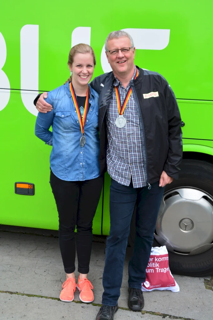 Ein Mann und eine Frau posieren lächelnd vor einem grünen Bus, beide tragen Medaillen um den Hals. Die Frau in einem Jeanshemd und mit orangenen Sportschuhen steht links, während der Mann in einem karierten Hemd und einer schwarzen Jacke rechts steht.