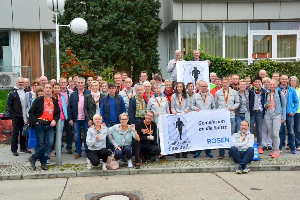 Gruppe von Menschen mit Medaillen posiert lächelnd für ein Gruppenfoto vor einem Gebäude, hält Banner der Lauffreunde Emsland.