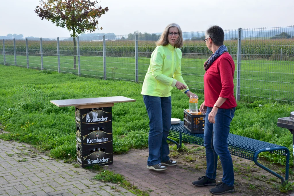 Zwei Frauen stehen im Freien, eine in einem leuchtend gelben Oberteil und die andere in einem roten Shirt. Sie interagieren an einem Tisch aus Krombacher-Kisten, während im Hintergrund eine Wiese und ein Zaun sichtbar sind.