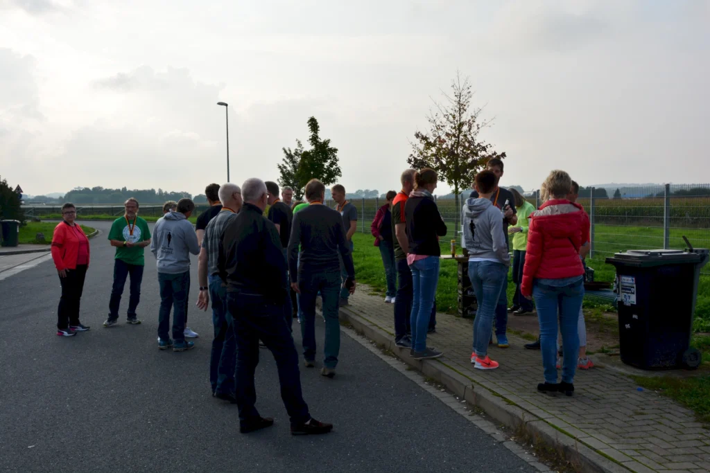 Gruppe von Menschen, die an einer Straße stehen und sich unterhalten, während sie an einem Event teilnehmen. Im Hintergrund sind Bäume und ein Feld zu sehen, sowie Mülltonnen und eine Straßenlaterne.