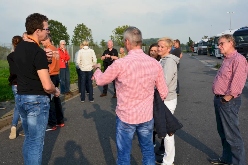 Gruppe von Menschen versammelt sich auf einem Parkplatz bei Tageslicht, umgeben von Lkw und grüner Landschaft.