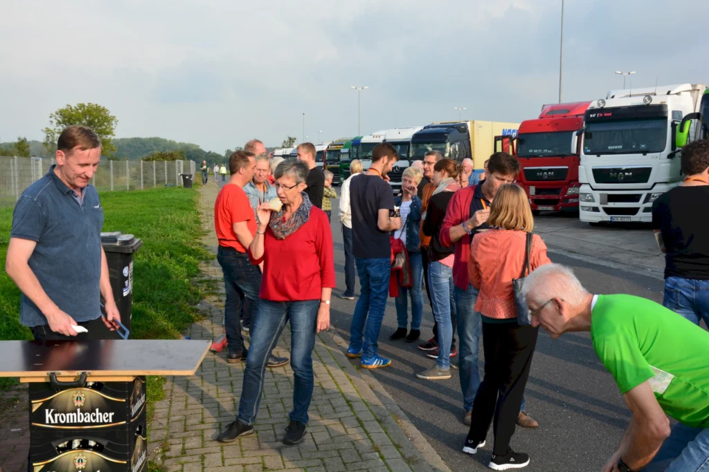 Menschenmenge bei einem Event an einem Parkplatz, mit Lkw im Hintergrund. Ein Mann steht an einem Tisch, während andere Gäste mit Getränken und Snacks interagieren. Die Teilnehmer tragen farbige Kleidung und scheinen sich zu unterhalten und zu genießen.